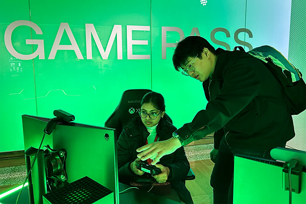 Two people looking at a computer: one is a women who is sitting holding a gaming controller and the other person is a man standing pointing to the screen