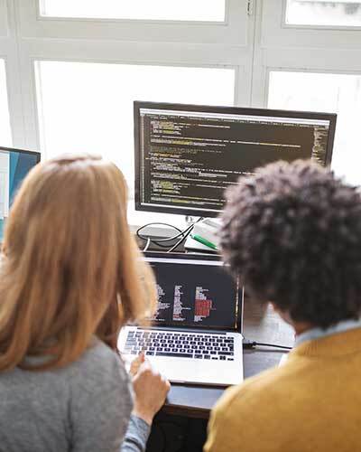 Over-the-shoulder view of two people working at a desk with multiple monitors displaying lines of computer code.